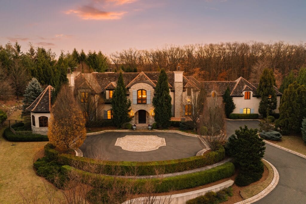 A stunning aerial view of a luxury estate at twilight, featuring a grand circular driveway, meticulously landscaped greenery, and warm interior lighting that highlights the home's elegant stone façade, set against a backdrop of trees and a soft sunset sky.