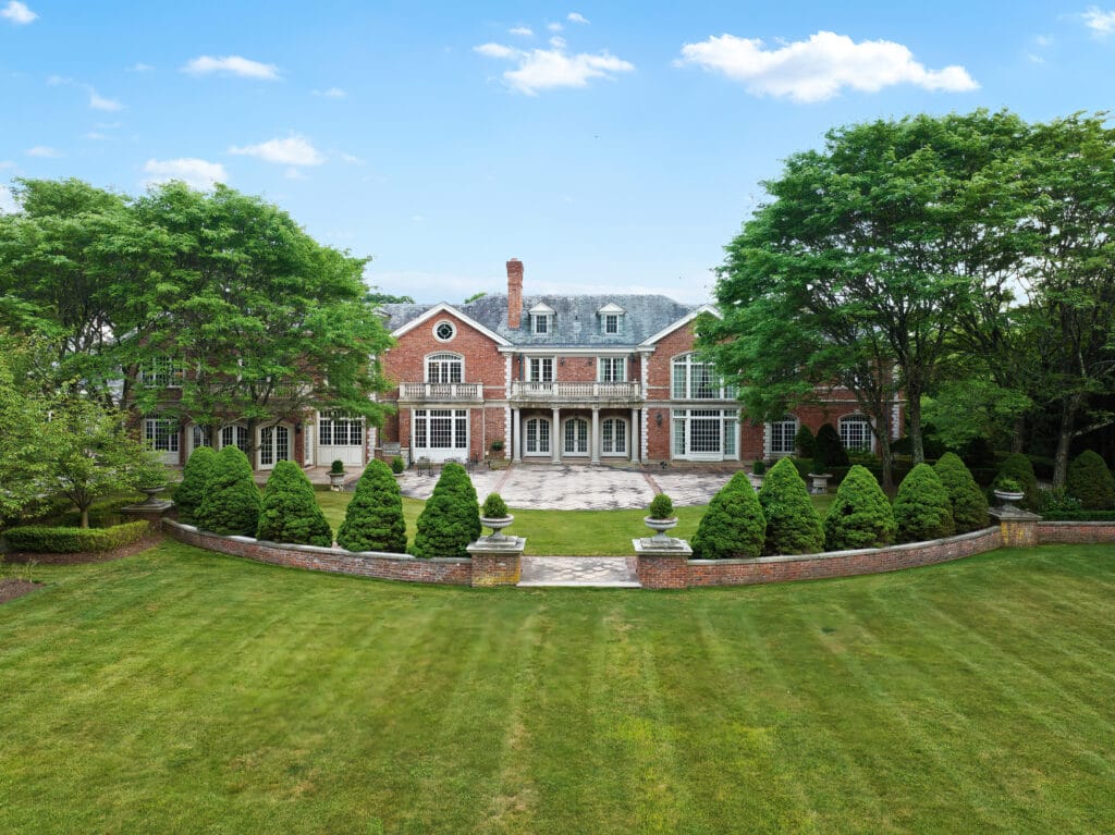 A breathtaking aerial view of a stately brick manor surrounded by lush greenery and perfectly manicured lawns, with symmetrical landscaping leading to an elegant patio area, set under a bright blue sky with scattered clouds.